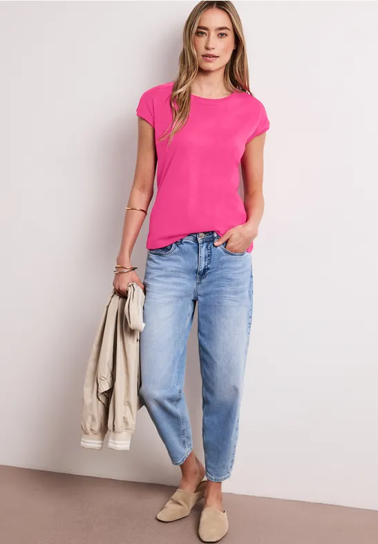 Een vrouw draagt een roze t-shirt, lichtblauwe jeans, houdt een beige jas vast en heeft beige schoenen aan.