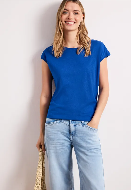 Vrouw in blauw t-shirt en lichtgekleurde spijkerbroek, hand in zak, met strotas tegen witte achtergrond.