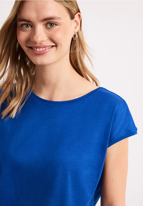 Een vrouw draagt een blauw T-shirt met korte mouwen, heeft lang haar en oorbellen, gezicht onscherp.