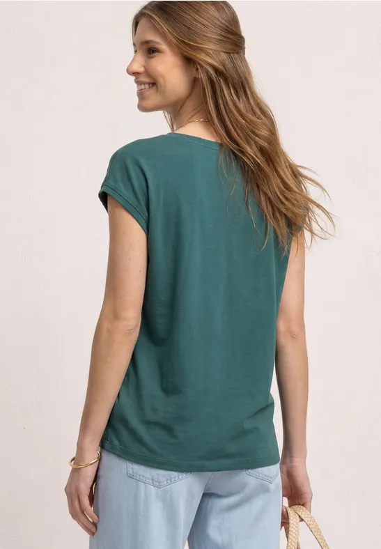 Vrouw van achteren in een groen shirt en spijkerbroek voor een lichte, neutrale achtergrond.