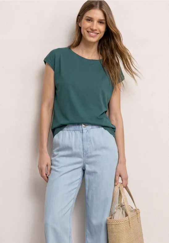 Vrouw met groen T-shirt en lichtblauwe jeans, met strooien tas, tegen een lichte achtergrond.