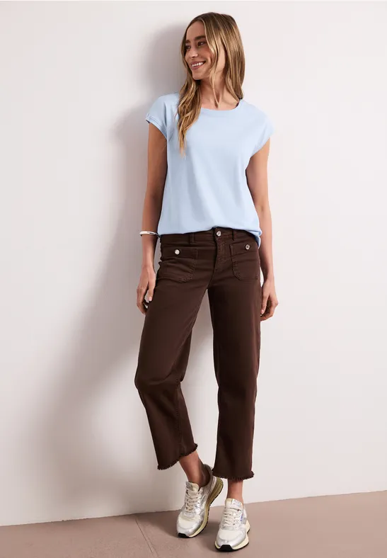 Vrouw in licht t-shirt, bruine 3/4-broek en sneakers voor een neutrale achtergrond.