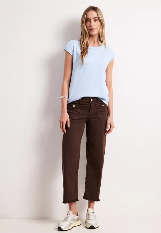 Vrouw in lichtblauw shirt, bruine broek en witte sneakers voor een witte muur.