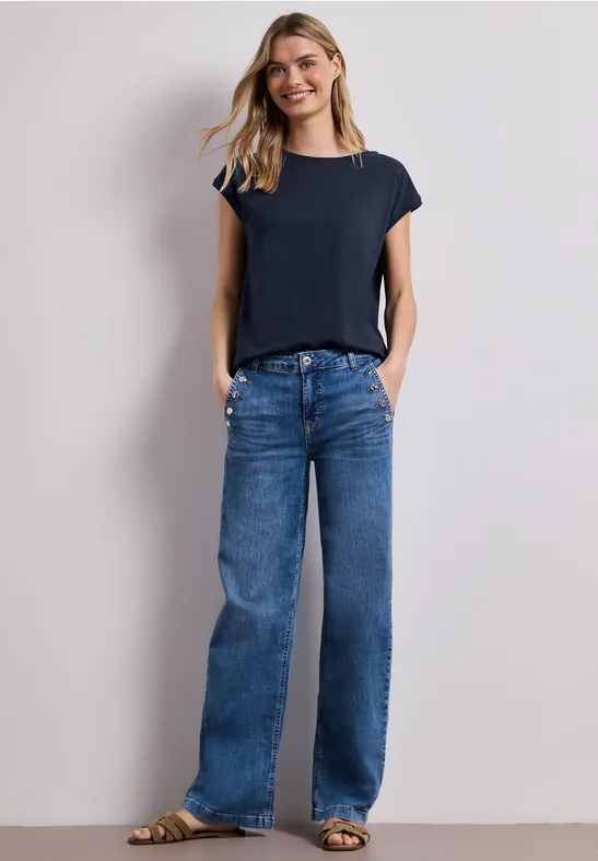 Vrouw in marineblauw T-shirt en wijdvallende jeans tegen witte muur.