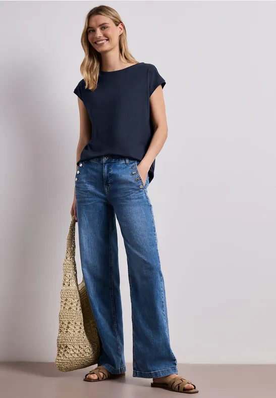 Vrouw in marineblauw shirt, wijde jeans, strooien tas en sandalen tegen neutrale achtergrond.