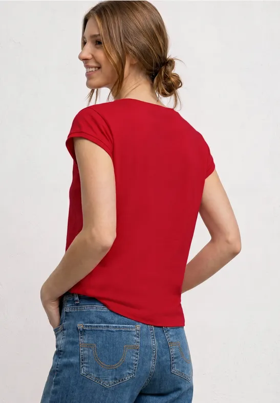 Vrouw van achteren in een rood t-shirt en blauwe jeans, handen in de zakken, haar in een lage knot.