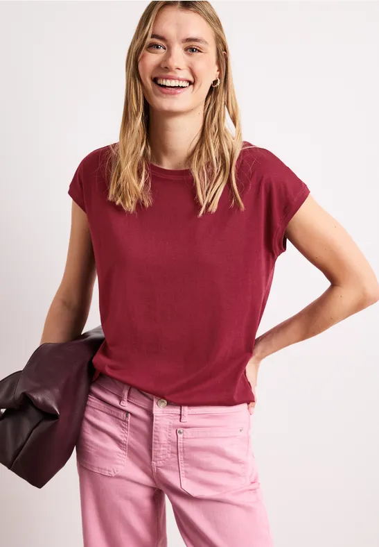 Vrouw in bordeaux t-shirt en roze jeans, houdt een tas vast voor een lichte achtergrond.