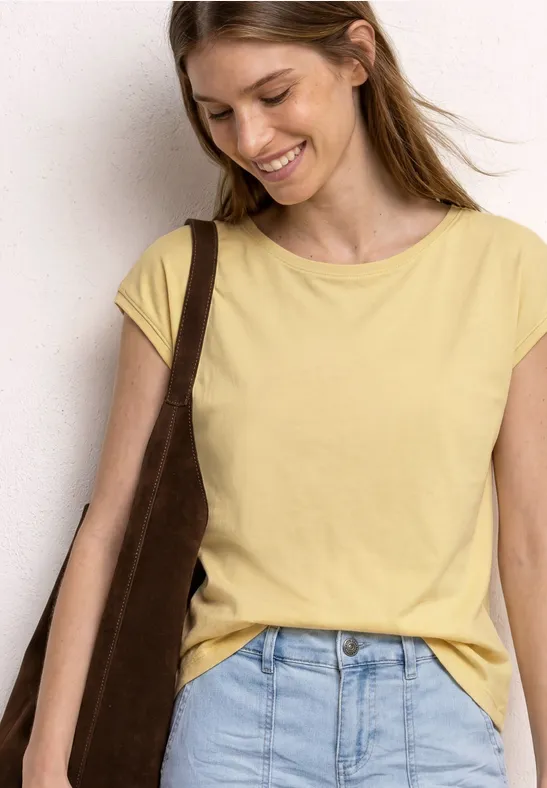 Frau in gelbem T-Shirt und hellblauen Jeans mit brauner Schultertasche vor neutralem Hintergrund.