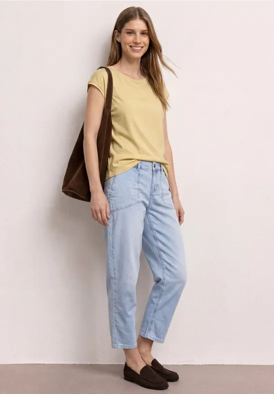 Frau in gelbem T-Shirt, hellblauen Jeans, braunen Loafern und brauner Tasche vor hellem Hintergrund.