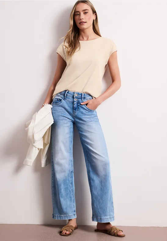 Vrouw leunt tegen muur in beige T-shirt en lichte, wijde jeans, houdt een jas in haar hand.