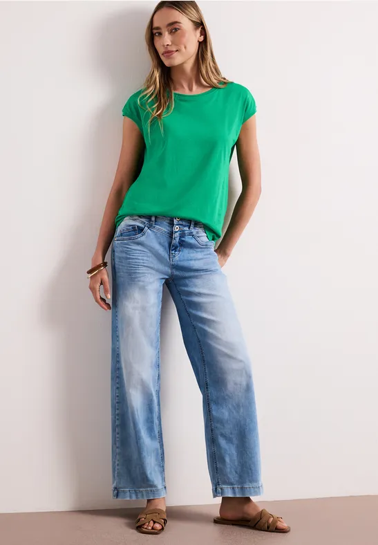 Vrouw draagt groen T-shirt en wijde lichtblauwe jeans, ontspannen poserend voor een witte muur.