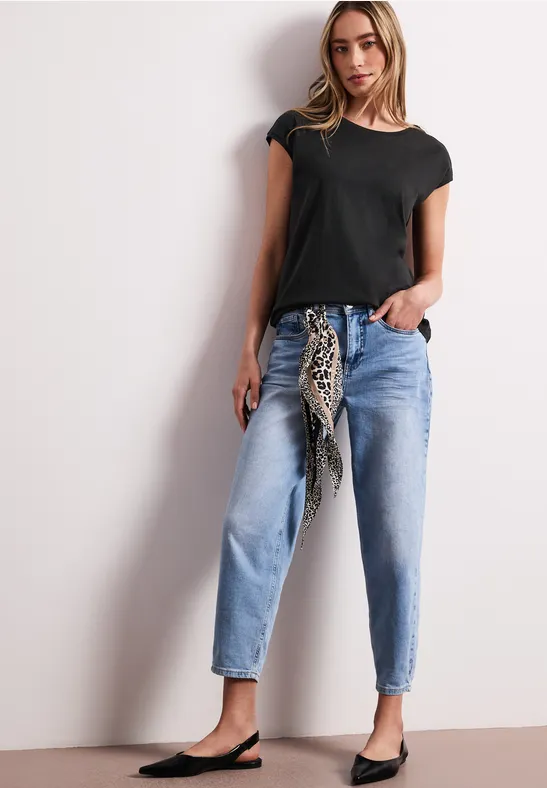 Vrouw in zwart T-shirt, lichtblauwe jeans, met een knoop-sjaal en platte schoenen.