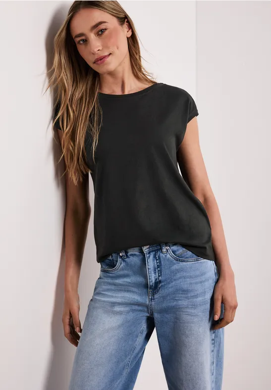 Een vrouw draagt een zwart t-shirt en lichtblauwe jeans, staat ontspannen voor een lichte achtergrond.