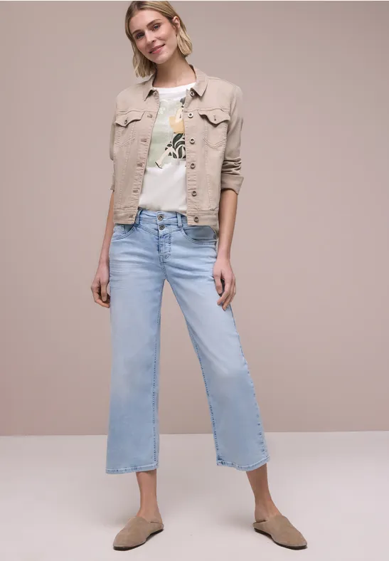Vrouw met lichtblauwe rechte jeans, wit T-shirt, beige jack en beige instappers tegen neutrale achtergrond.