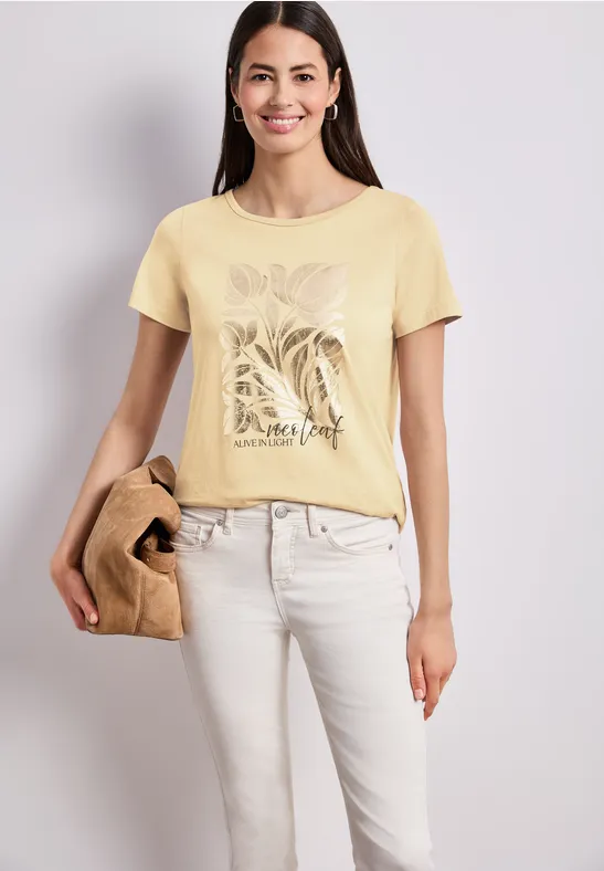 Vrouw draagt lichtgeel T-shirt met plantenprint en witte broek, houdt een tas vast voor een neutrale achtergrond.