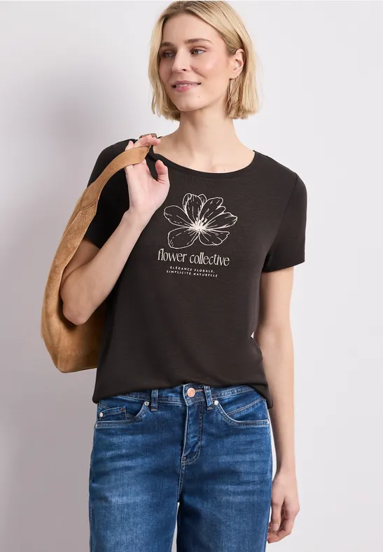 Vrouw draagt zwart T-shirt met witte lotusbloem en tekst, een spijkerbroek en een schoudertas.