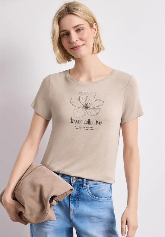 Frau in beigem T-Shirt mit Blumenmotiv und Jeans, hält passende Jacke in der Hand.