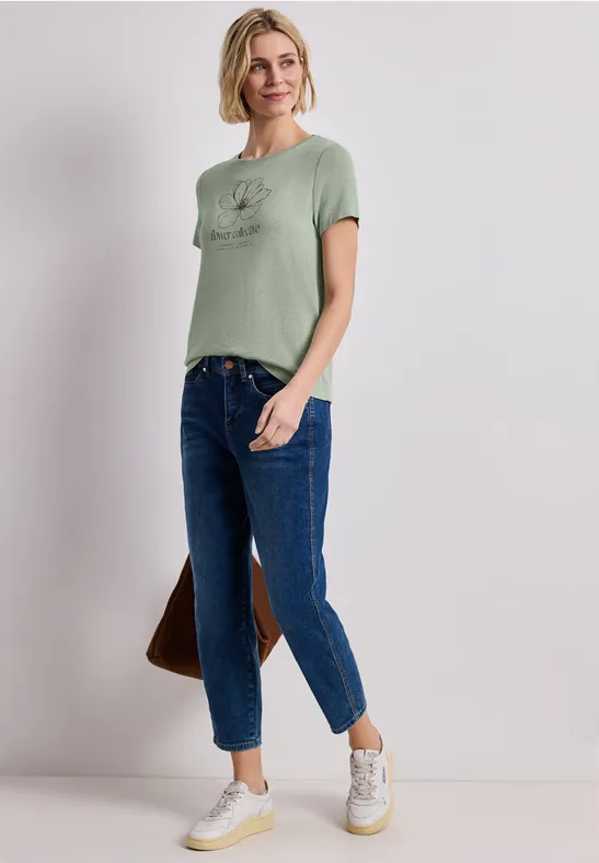 Frau in grünem T-Shirt, blauen Jeans, weißen Sneakern, steht lässig, trägt braune Tasche.