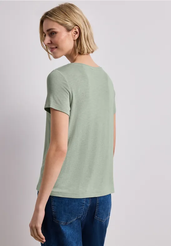 Frau mit kurzem blonden Haar trägt ein hellgrünes T-Shirt und blaue Jeans, von hinten fotografiert.