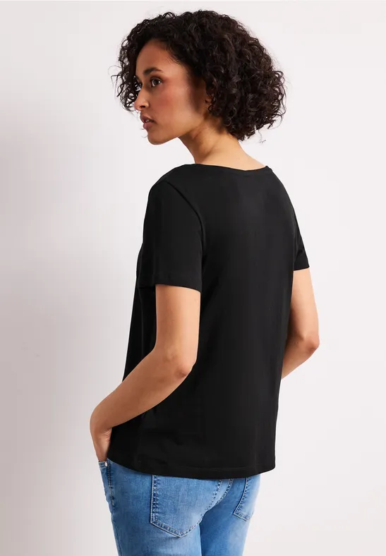 Vrouw in profiel met zwart t-shirt en blauwe spijkerbroek, handen in zakken, krullend haar.