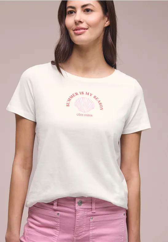 Vrouw met wit t-shirt met rode opdruk en roze broek voor een lichte achtergrond.
