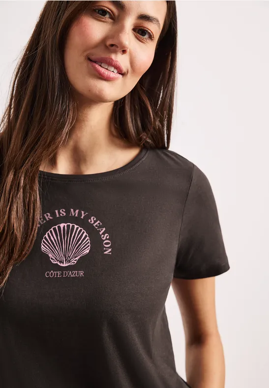 Zbliżenie kobiety w czarnej koszulce z nadrukiem muszli i napisem 'Sea is my season'.