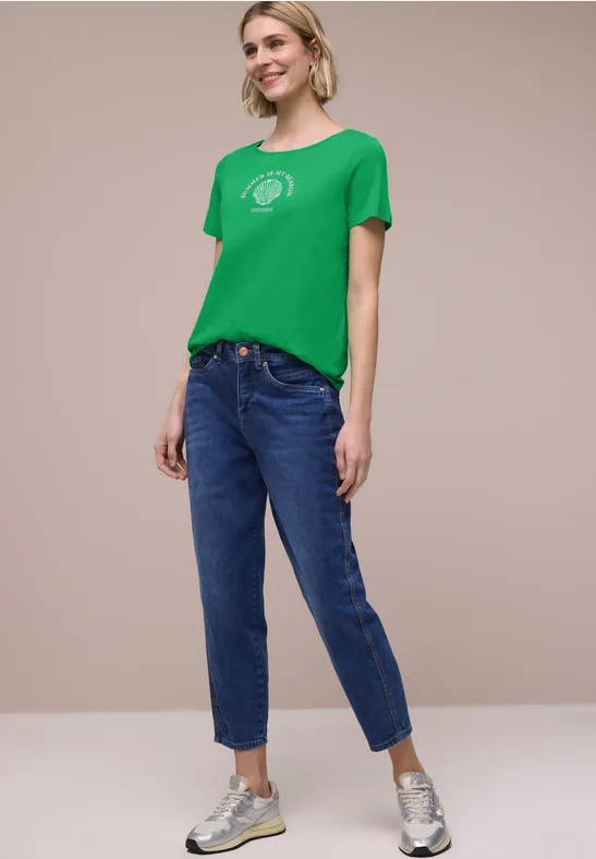 Vrouw met groen T-shirt, blauwe spijkerbroek en zilverkleurige sneakers voor een neutrale achtergrond