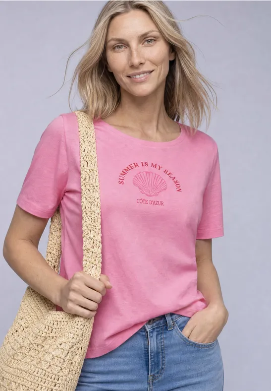 Frau mit pinkem T-Shirt, Jeans und geflochtener Tasche lächelt in die Kamera.