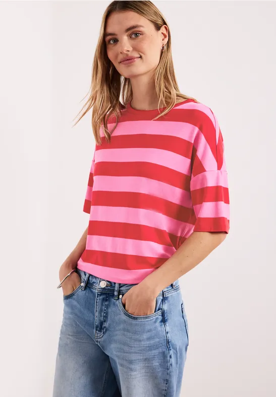 Vrouw in roze-rood gestreept t-shirt en blauwe spijkerbroek, nonchalant pose met handen in haar zakken.