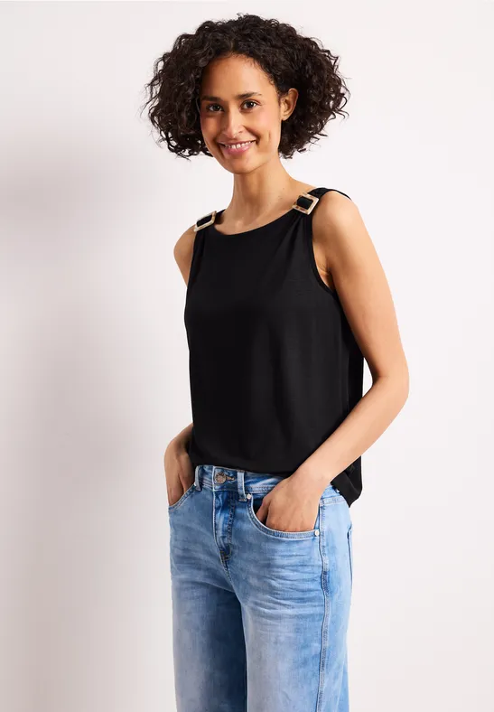 Een vrouw in een zwart spaghettitop en blauwe jeans tegen lichte achtergrond.