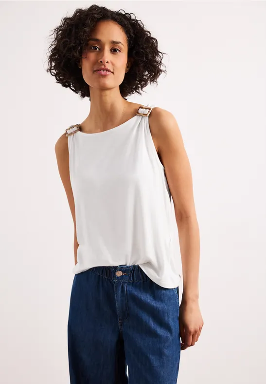 Vrouw met krullend haar in een wit mouwloos topje en blauwe jeans voor een lichte achtergrond.