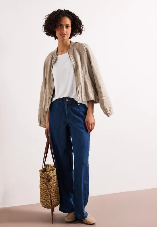 Vrouw in wit T-shirt, beige jasje, wijde jeans, gevlochten rieten tas en beige loafers.