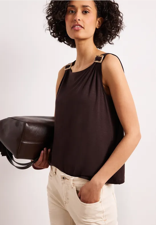 Femme en haut marron sans manches à bretelles dorées, pantalon clair, tenant un grand sac en cuir marron.