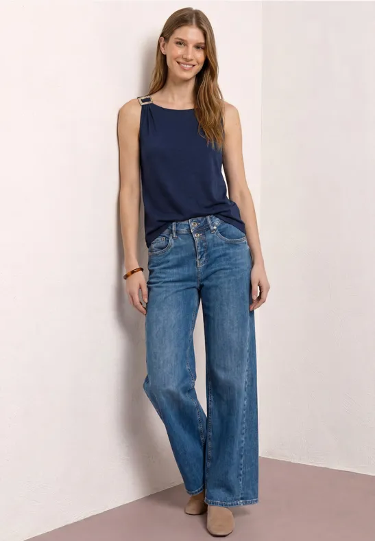 Vrouw leunt tegen muur, donkerblauw hemdje, wijde jeans en beige enkellaarsjes voor een strakke achtergrond.