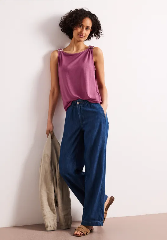 Vrouw met krullend haar in een paarse top en wijde jeans, met een beige jasje in haar hand.