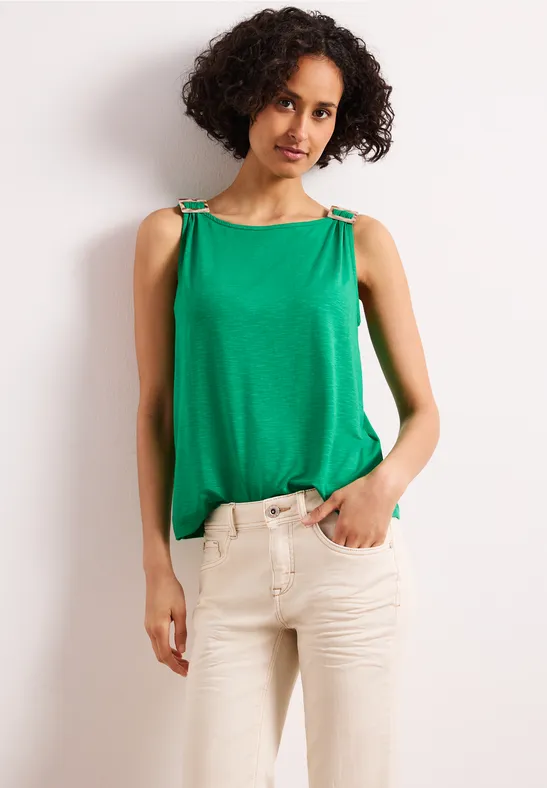 Vrouw met krullend haar draagt een mouwloze groene top met sierbanden en een beige broek.