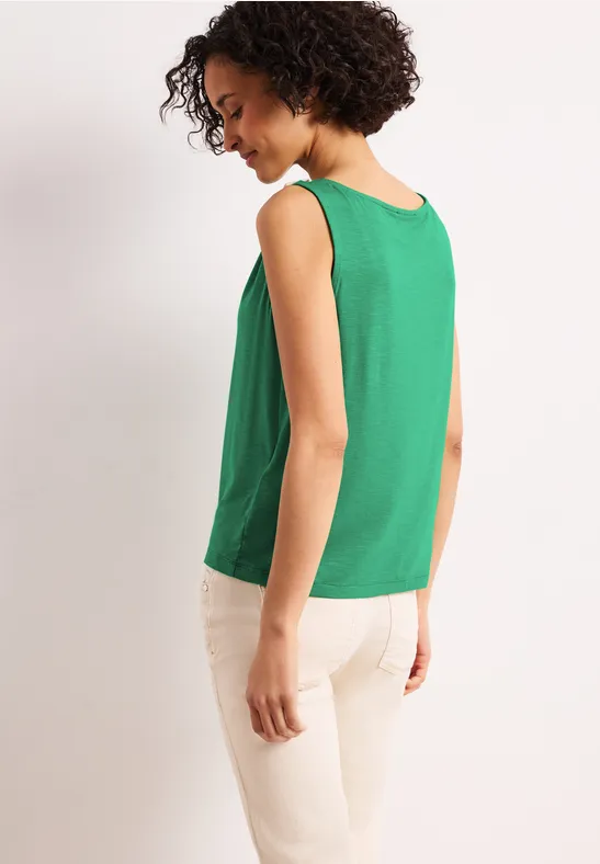 Vrouw van achteren in een groen mouwloos topje en beige broek voor een lichte achtergrond.