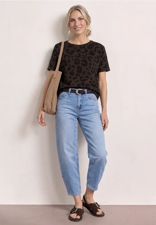 Persoon voor witte muur draagt zwart leopard-T-shirt, lichte jeans, zwarte sandalen en katoenen tas.