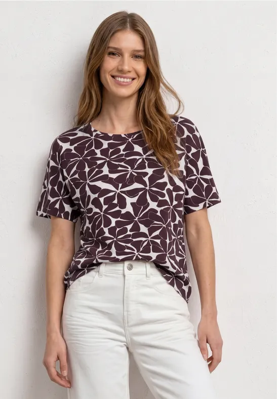 Vrouw in paars-wit t-shirt met patroon en lichte broek voor een neutrale achtergrond.