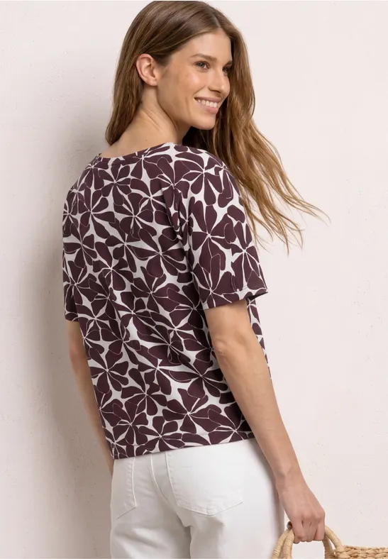 Vrouw van achteren in bruin-witte bladprint top, witte broek en met een strooien tas.