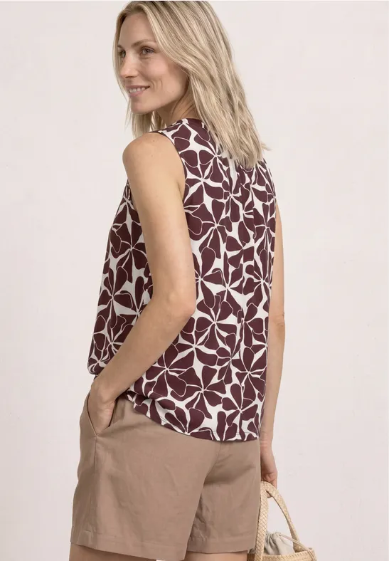 Ärmellose Bluse mit weiß-bordeauxfarbenem Blattmuster, beigen Shorts und Strohtasche.