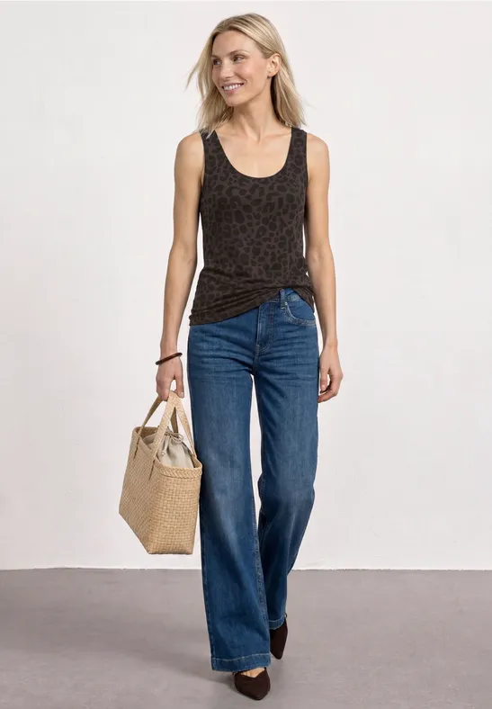 Een vrouw draagt een zwart tanktop met patroon, wijde blauwe jeans en houdt een strooien tas vast.