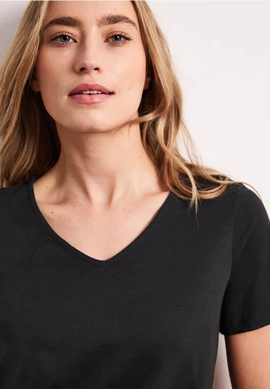 Portret van een jonge vrouw in zwart V-hals T-shirt, golvend haar, lichte achtergrond.