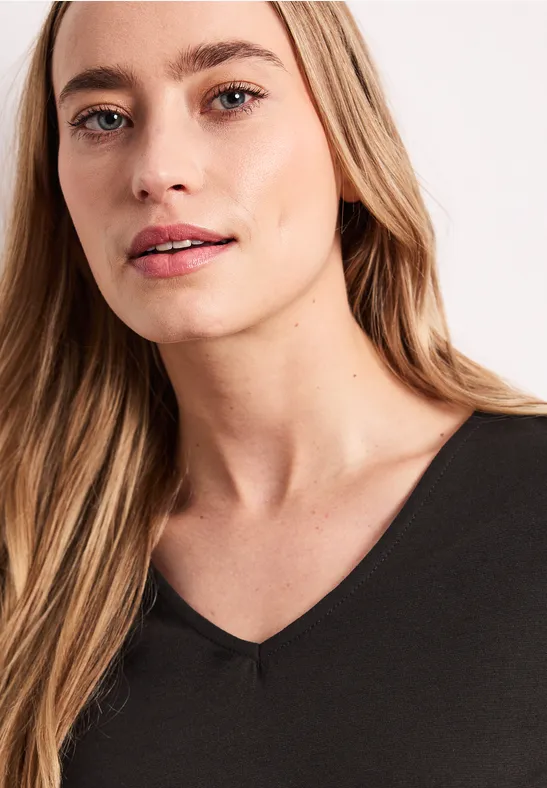 Femme aux cheveux longs portant un t-shirt noir à col V sur fond clair.