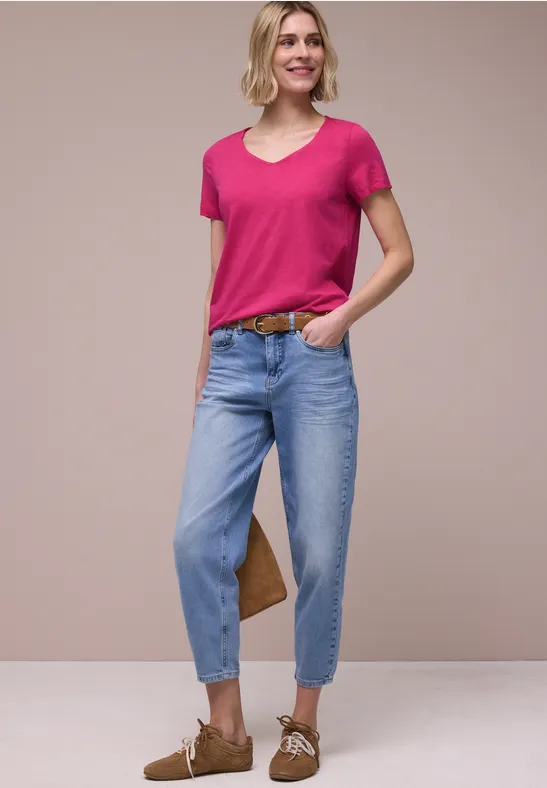 Persoon in roze T-shirt, lichtblauwe omgeslagen jeans, bruine riem en bruine veterschoenen voor beige achtergrond.