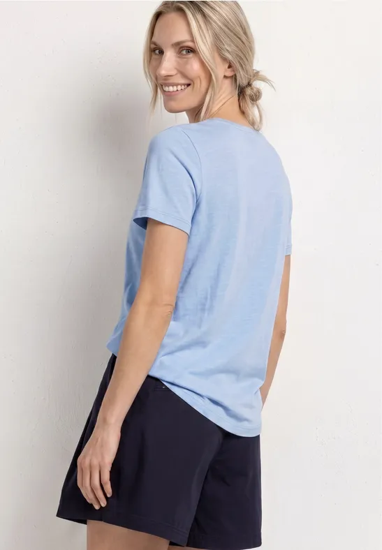 Frau steht seitlich, trägt hellblaues T-Shirt und schwarze Shorts vor weißem Hintergrund.