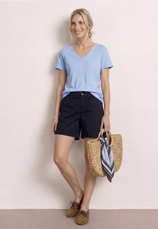 Frau im hellblauen T-Shirt, dunkelblauen Shorts und Basttasche mit Tuch, entspanntes Sommeroutfit.