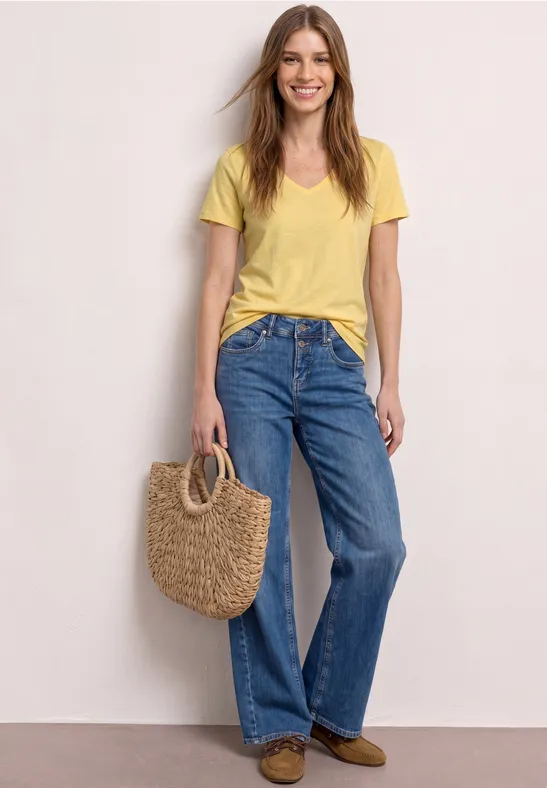 Frau in gelbem T-Shirt und weiten Jeans, hält Basttasche, steht vor hellem Hintergrund.