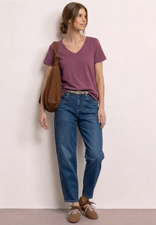 Vrouw leunt tegen muur, draagt paars T-shirt, blauwe high-waist jeans, bruine schoenen en tas.