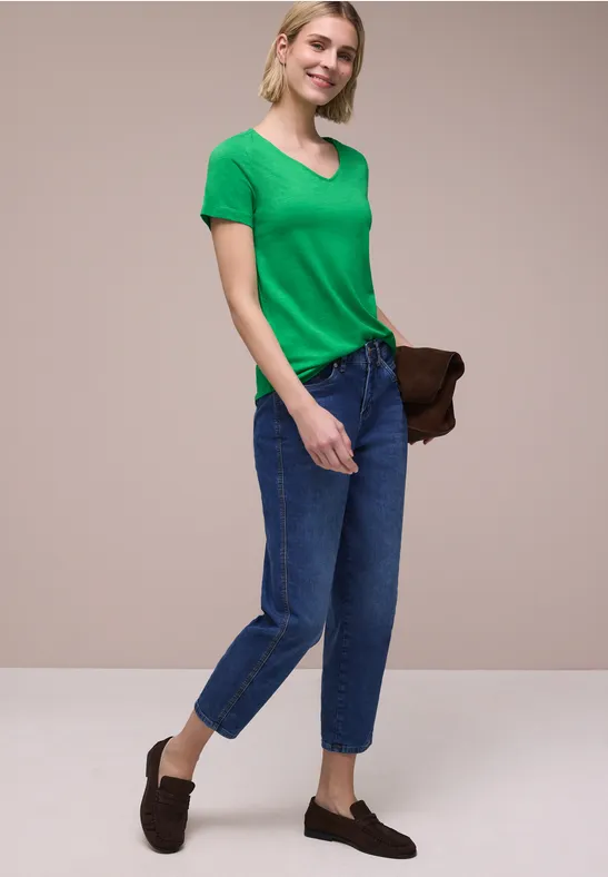 Vrouw met groen T-shirt, blauwe spijkerbroek en loafers, houdt een bruine clutch vast.